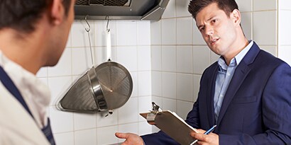 A long, narrow commercial kitchen with stainless steel finishes has several chefs in white busily preparing food.
