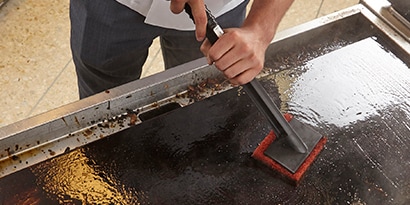 Una persona frotando una plancha con la Fibra de Limpieza Rápida para Planchas 3M™ Scotch-Brite™ 746 y el Soporte para Fibras Multiusos Scotch-Brite™ 405-R.

