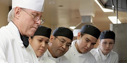 Un chef demuestra una técnica de preparación de alimentos a cuatro trabajadores de cocina que observan.
