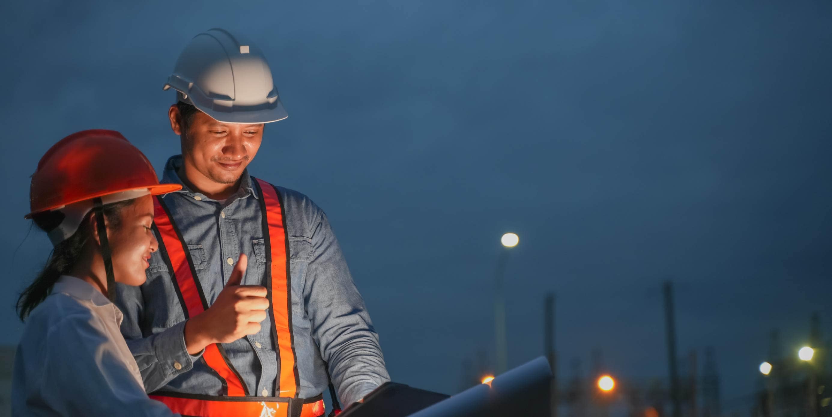 Personal de obra con cascos y chalecos reflectantes revisa planos al anochecer. 
