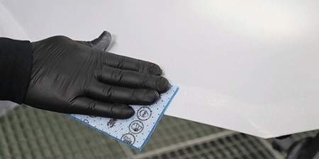 Una persona que lleva un guante negro utiliza una almohadilla abrasiva azul para lijar el borde de una superficie lisa y blanca.
