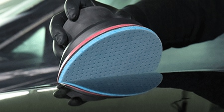 Una persona con guantes negros sostiene una lijadora eléctrica con un disco abrasivo flexible de espuma 3M™ Hookit™ azul, colocada sobre una superficie brillante, lista para lijar.
