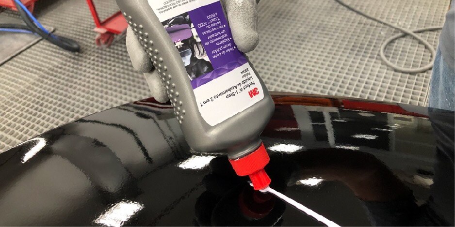 Persona aplicando un compuesto de pulido 3M sobre una superficie automotriz negra y brillante dentro de un área de taller.
