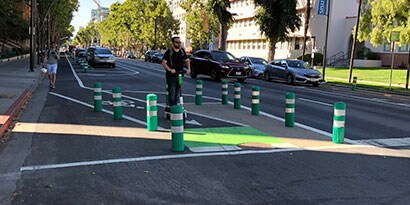 Urban road with person on scooter in protected bike lane, cars on road and pedestrian on road near sidewalk.
