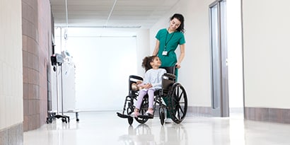 Una mujer llevando a una niña en silla de ruedas en un hospital.
