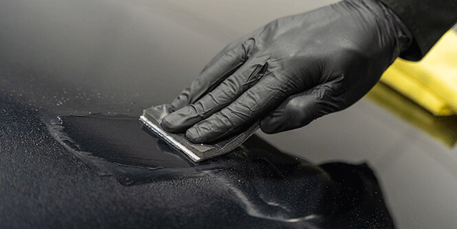 Una persona con guantes negros está aplicando un revestimiento a una superficie con un pequeño aplicador rectangular y esparce un líquido uniformemente sobre el área.
