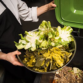 A restaurant worker lifts the lid of a bin to dispose of vegetables.