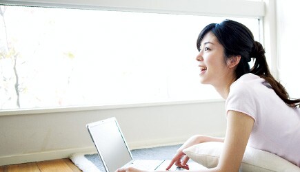 A young woman happily works on a laptop near a sunny window, unconcerned about glare.