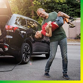 Hombre jugando con su hijo junto a un auto negro cargando en el camino de entrada.