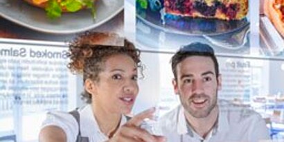 Una mujer y un hombre sonrientes miran un menú flotante y colorido en un restaurante.
